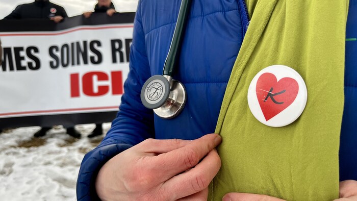 Un médecin arbore un macaron illustré d'un coeur et d'un K pour Kamouraska, devant une affiche indiquant Mes soins restent ici. 