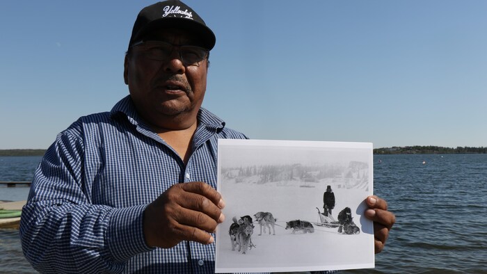 Un homme portant une casquette montre une vieille photo en noir et blanc d'un équipage de chiens de traîneau.