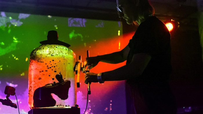 L'artiste est debout, devant un bocal avec un fluide magnétique. Il y a des éclairages colorés.