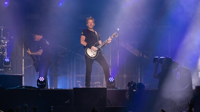Le chanteur de Nickelback debout sur scène, guitare à la main, dans une lumière violacée.