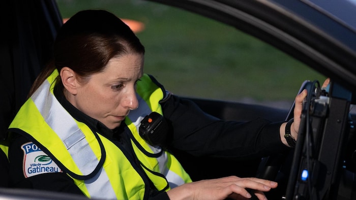 L'agente Andrée East à bord d'une voiture de police en uniforme. Elle consulte l'écran tactile situé à la droite de son volant.