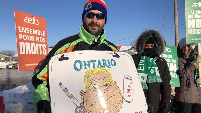 Un homme pose avec une pancarte où l’on peut voir une caricature de Doug Ford.
