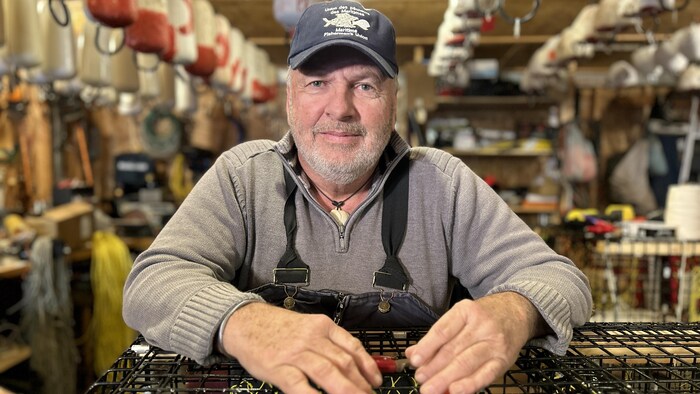 Réjean Comeau porte une casquette de l'Union des pêcheurs des Maritimes ainsi qu'une salopette.  Il est debout dans lad   garage, accoté contre un casier.