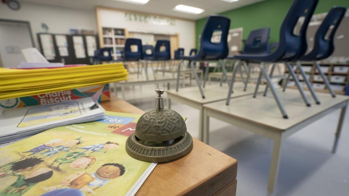 Cloche d'école ancienne et des cahiers sur un bureau en bois, avec des chaises empilées sur des tables à l'arrière-plan d'une salle de classe primaire vide.