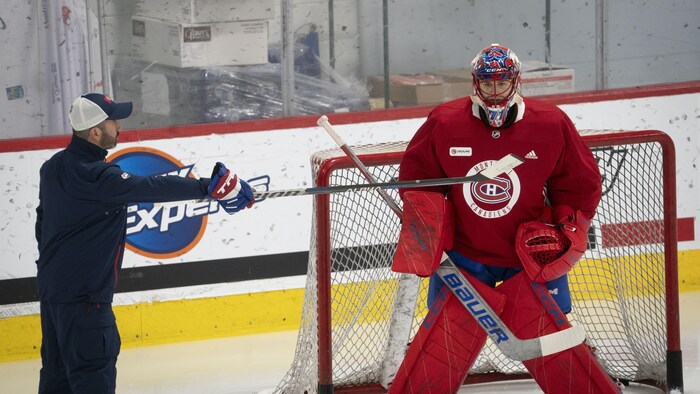 Un gardien de but au hockey posté devant son filet écoute les conseils d'un entraîneur qui se tient près de lui.