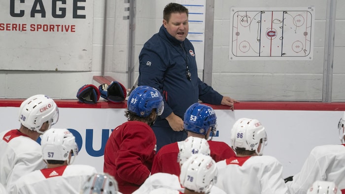 Un entraîneur de hockey parle à ses joueurs devant un tableau, en bordure de la patinoire.