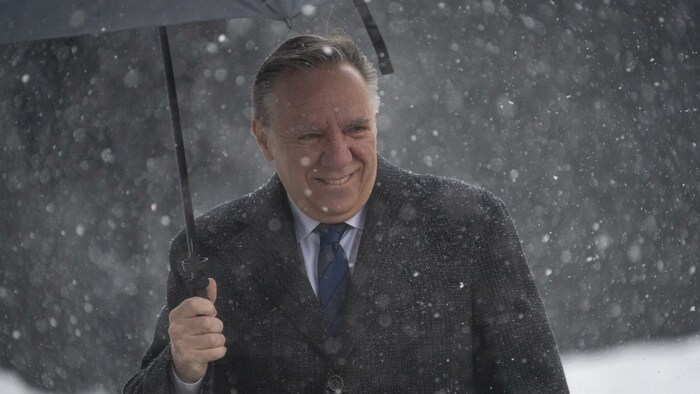 François Legault se tient dehors sous la neige avec un parapluie.