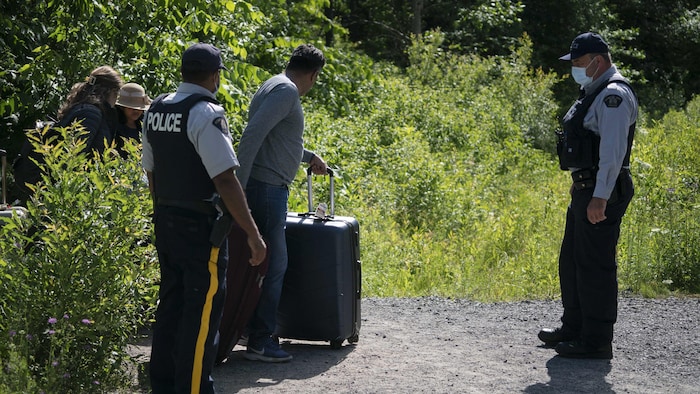 Deux agents frontaliers encadrent un homme tirant une valise.