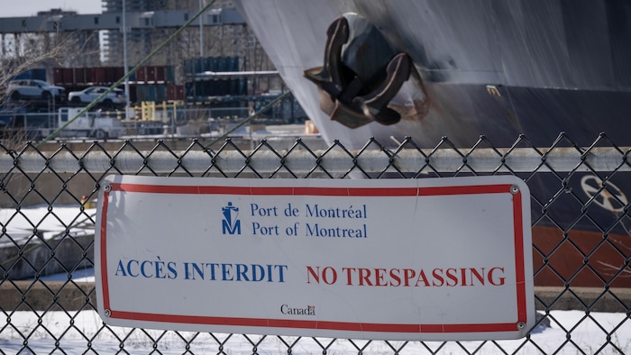 Une pancarte «Port de Montréal - Accès interdit - No trespassing» sur une clôture derrière laquelle on aperçoit l'ancre d'un bateau et, au loin, une remorque transportant des voitures.