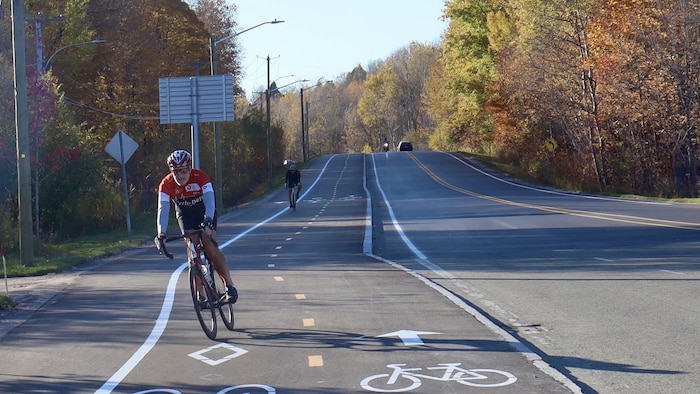Un cycliste casqué roule sur une voie réservée asphaltée longeant une route bordée d'arbres d'automne.