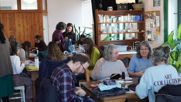 Des personnes réunies autour de tables sur lesquelles il y a des vêtements, de boutons, de la laine, du tissu.
