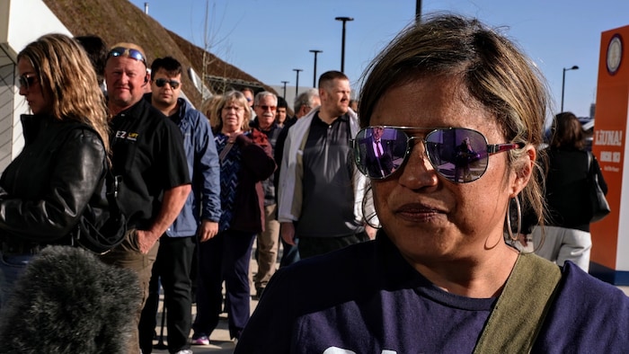 Une femme portant des lunettes fumées accorde une entrevue devant un groupe de gens.