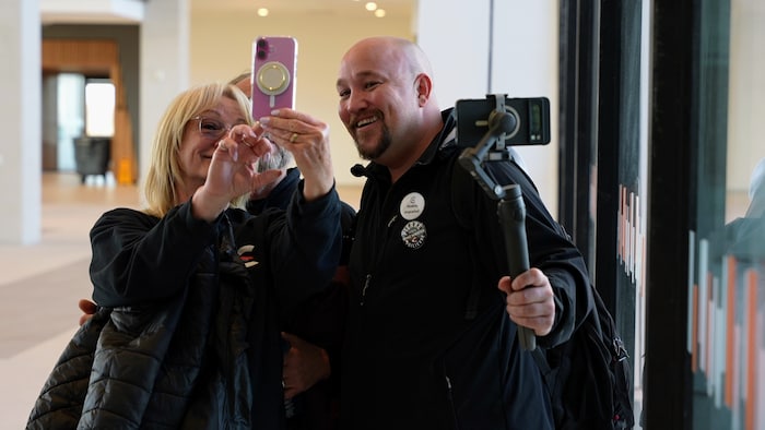 Une femme blonde et un homme chauve se tiennent côte à côte et sourient devant un téléphone cellulaire determination prendre un égoportrait.