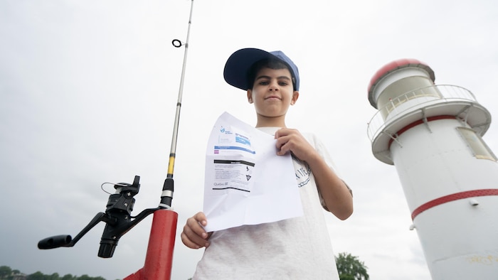 Massyle exhibe son permis, avec sa canne à pêche et le phare de Lachine en arrière-plan.