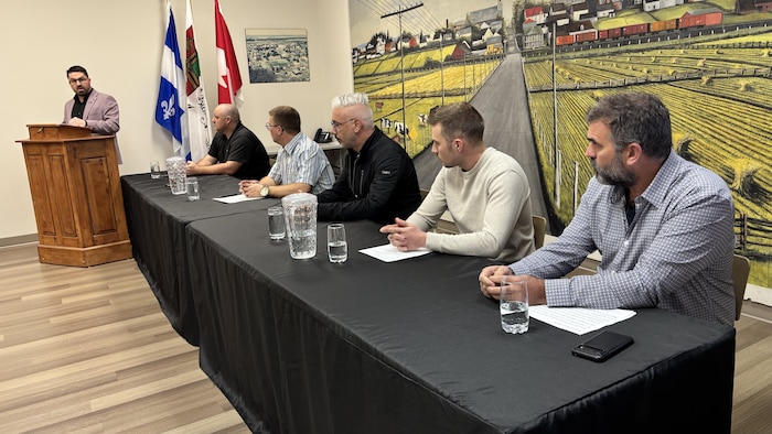 UN groupe de cinq personnes sont assises à une table de conférence avec le maire qui parle au lutrin.