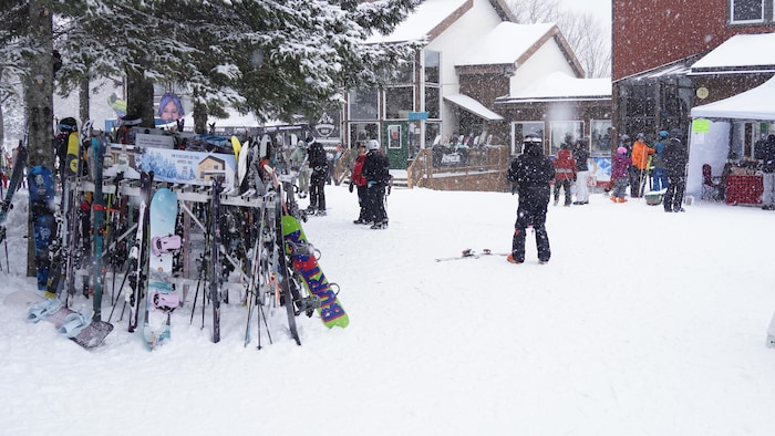 À gauche, des ski et des planches à neige rangés sous un arbre enneigé, à droite, plusieurs personnes en tenue d'hiver se déplacent au pied du Mont-Comi.