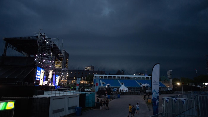 Overview of the plains of Abraham in Quebec under a very dark and stormy sky. In the foreground on the left. a large concert scene is illuminated with blue and white lights, with visible metal scaffolding. national day show quebec: public