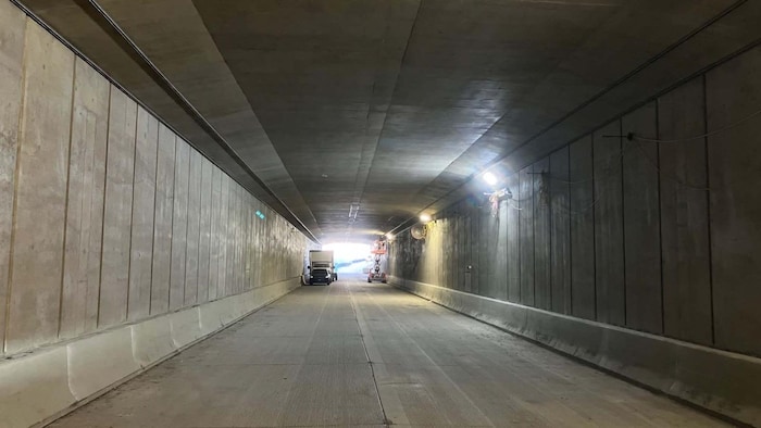 Intérieur d'un tunnel en béton éclairé où stationnent des véhicules de chantier près de la sortie lumineuse.
