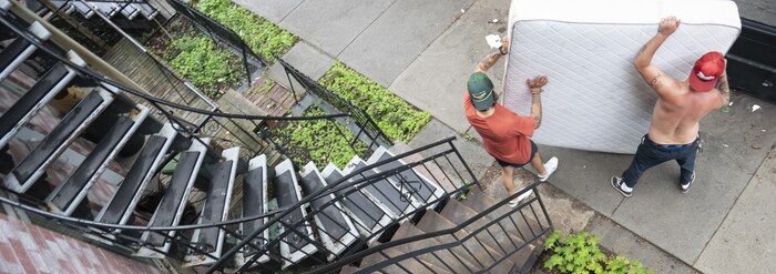 Deux hommes portant une casquette, dont un torse nu, manipulent un matelas sur un trottoir en bas d'un escalier extérieur.