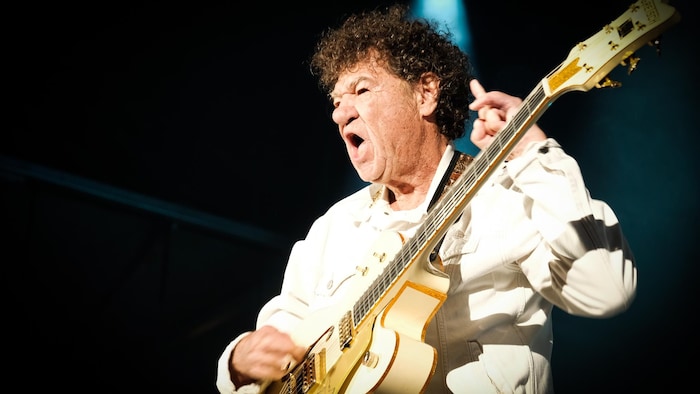 Robert Charlebois tient une guitare et a une main levée.
