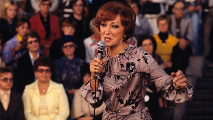 Lucille Dumont vêtue d'une robe fleurie, tenant un micro et chantant au Complexe Desjardins en 1977; en arrière plan flou, une foule assise