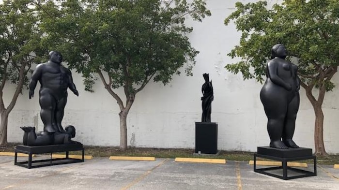 Extérieur. Jour. Trois personnages sculptés par Fernando Botero se tiennent debout dans un stationnement.