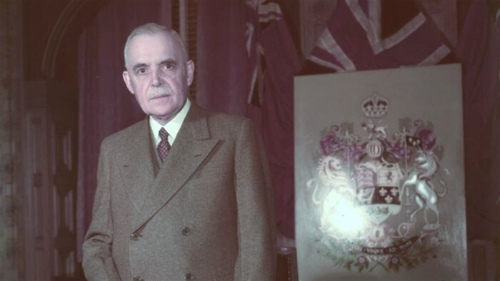 Louis St-Laurent pose debout à côté des armoiries du Canada.