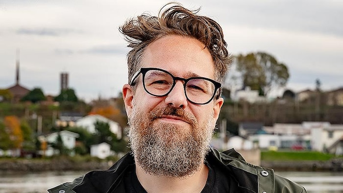 Un homme portant barbe, lunettes, t-shirt noir et veste verte, photographié avec la rivière et la ville de Matane à l'arrière-plan.