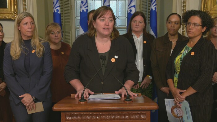 Une femme en conférence de presse entourée d'intervenantes et de drapeaux du Québec.