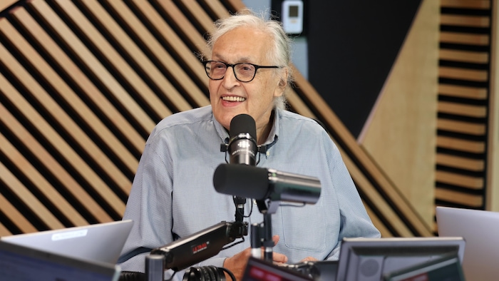 Il est assis en studio et sourit.