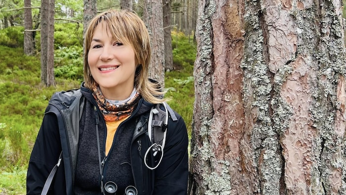 Andrée Gendron sourit, accotée sur un tronc d'arbre, en forêt, une paire de jumelles pendant à son cou.