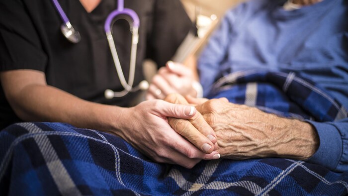 Une infirmière tient la main d'une aînée, qui est couchée dans un lit.