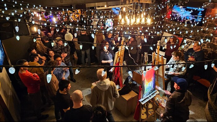 Une foule gravite autour d'artistes qui peignent des toiles en direct dans un café-bar.