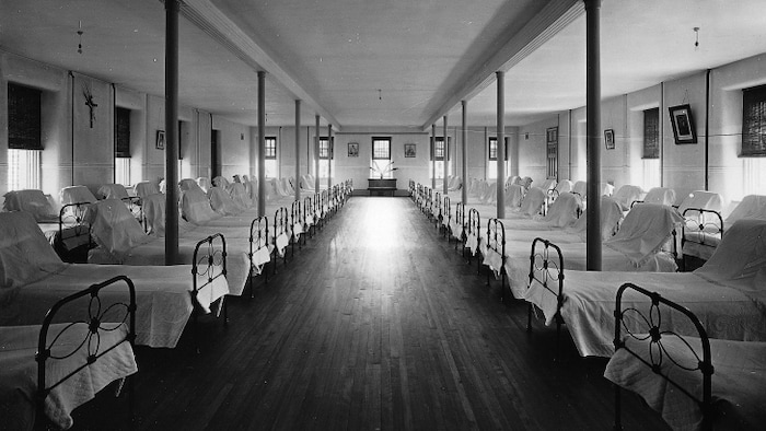 Photo en noir et blanc montrant plusieurs rangées de lits vides recouverts de draps blancs, et un crucifix sur le mur.