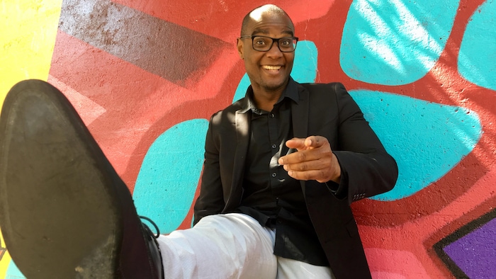 Trésor Otshudi adossé à un mur coloré a la jambe levée et pointe la caméra du doigt le sourire aux lèvres. 