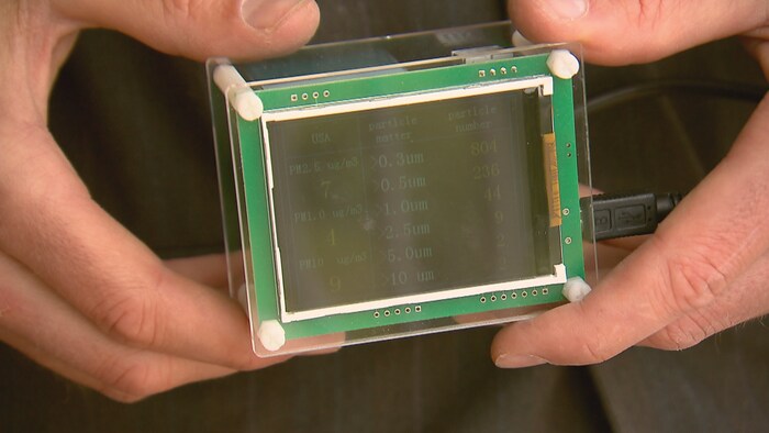 Des mains tiennent un boîtier avec un écran et des indications sur la qualité de l'air.