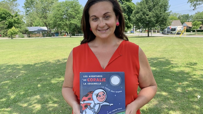 Celeste Kurcz de Windsor, souriante, est debout en plein air et tient dans ses mains le livre jeunesse qu'elle a écrit 'Les aventures de Coralie la coccinelle'.