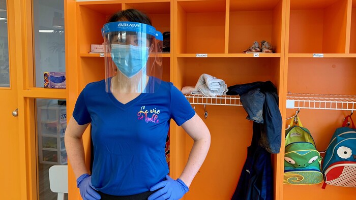 Une éducatrice avec des gants, un masque et une visière pose devant des casiers.