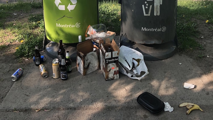 Des bouteilles de bière, un contenant en plastique, une peau de banane et deux caisses de bière au pied d'une poubelle de la Ville de Montréal.