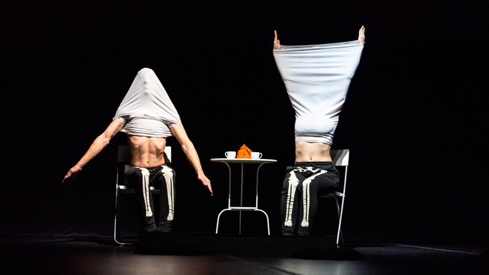 Deux artistes dont les pantalons évoquent un squelette, exécutent des mouvements des bras, attablés à une table. Leurs visages sont cachés par leurs chandails.