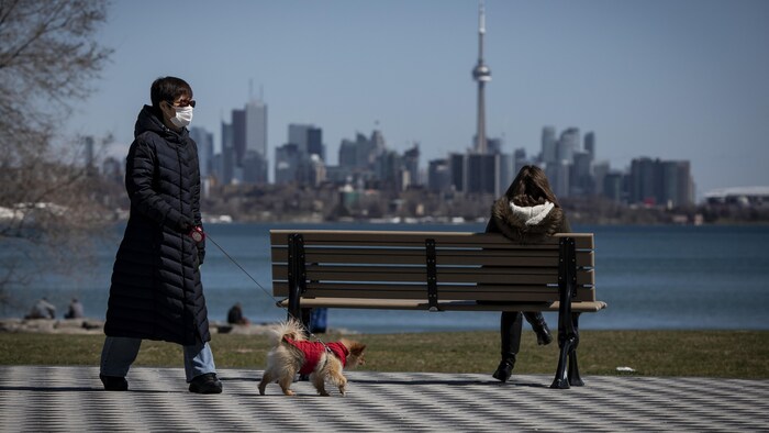 Une femme avec un masque passe à distance d'un banc public sur lequel une autre femme est assise. Cette dernière regarde la ville de Toronto.