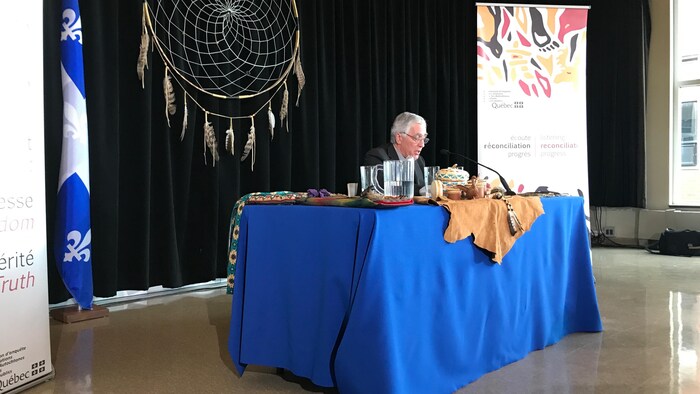 القاضي المتقاعد جاك فيان يقرأ توصيات وهو جالس إلى طاولة وخلفه علم كيبيك وستارة سوداء عُلق عليها أحد رموز السكان الأصليين.