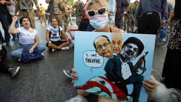 Une manifestante anti-gouvernementale libanaise tient une caricature représentant (de gauche à droite), le président du Liban, Michel Aoun, le président du Parlement, Nabih Berri, et le chef du mouvement chiite Hezbollah, Hassan Nasrallah, tous assis sur une personne drapée du drapeau national libanais, qui crie « Je ne peux plus respirer ».