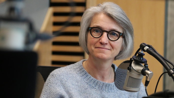 A woman smiles slightly as she listens intently behind a microphone in a radio studio. 