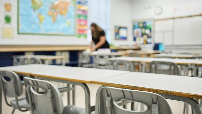 Des chaises et bureaux alignés dans une salle de classe d'une école, avec une enseignante travaillant à l'arrière-plan.