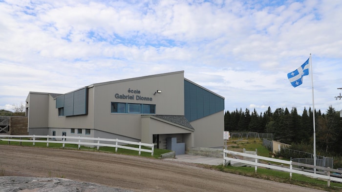 L'école Gabriel-Dionne, à Tête-à-la-Baleine, en Basse-Côte-Nord, vue de l'extérieur, en été avec un drapeau du Québec devant et la cour de récréation et des épinettes derrière.