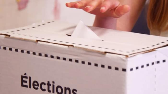 Gros plan d'une boîte de scrutin et d'une main qui glisse un bulletin de vote dans la boîte.
