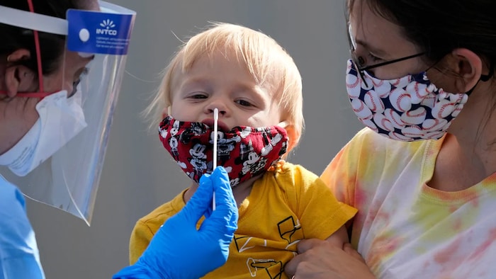 Une femme masquée porte dans ses bras un jeune enfant, auquel une infirmière est en train de faire un prélèvement de sécrétions nasales pour le dépistage de la COVID-19.