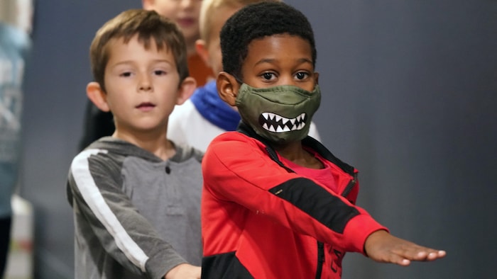 Quatre élèves, dont seul le premier porte un masque, marchent les uns à la suite des autres, le bras tendu, dans le couloir d'une école primaire.