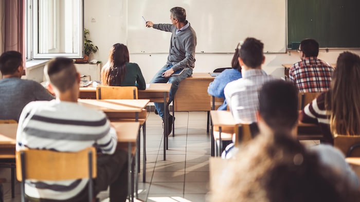 Un enseignant est assis sur son bureau et montre des choses au tableau. Des élèves sont assis en classe et écoutent.